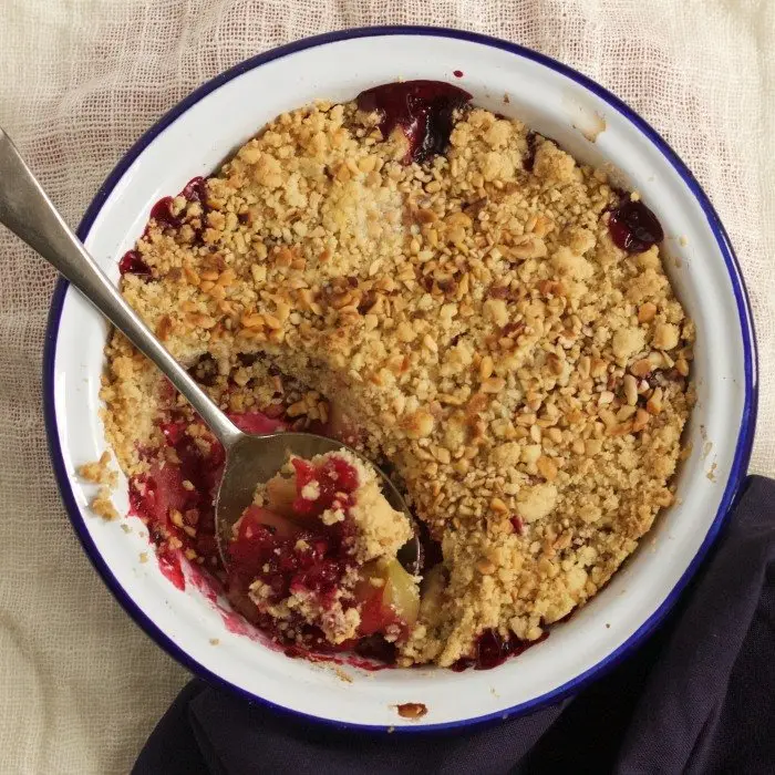 Crumble doré aux pommes et aux mûres dans un plat émaillé, une cuillère plantée